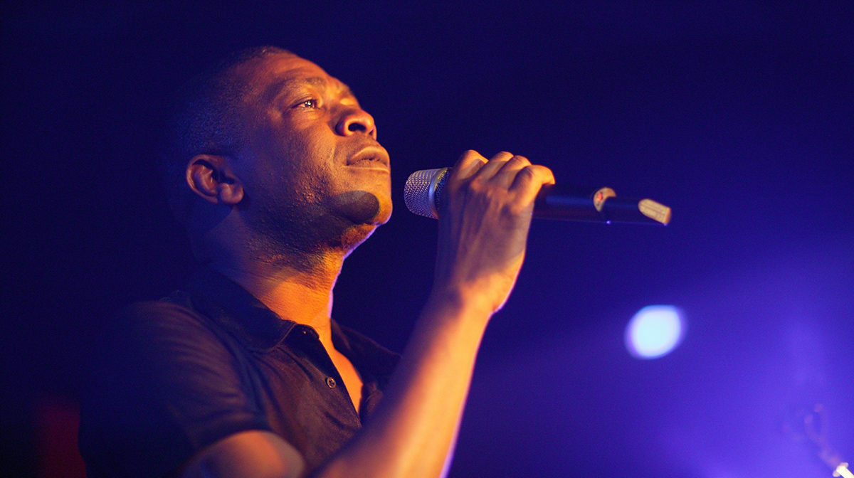 Youssou N’Dour sings passionately into a microphone against a navy backdrop.